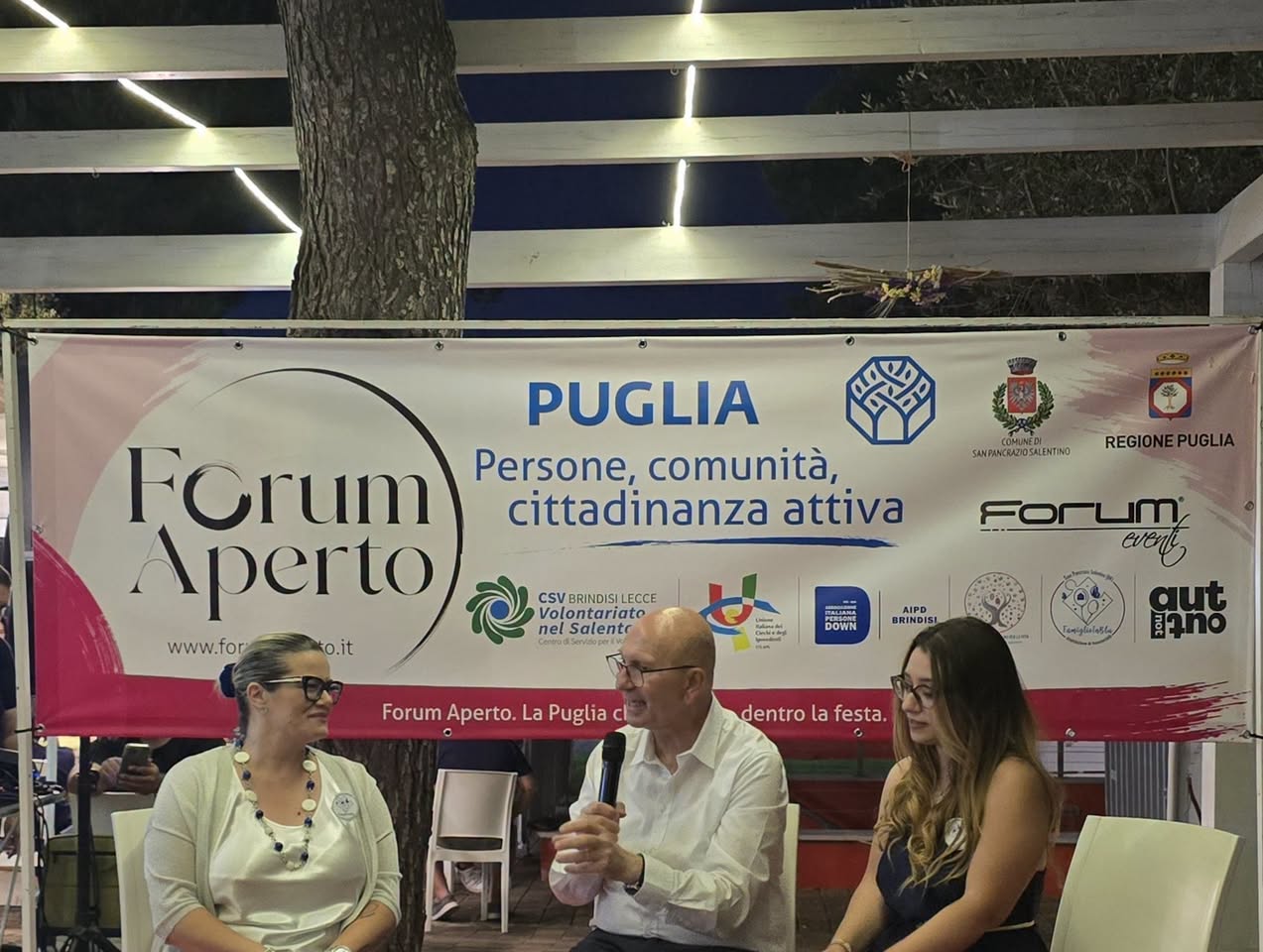 Un momento di dialogo al microfono davanti al banner di Forum Aperto con i loghi dei partner